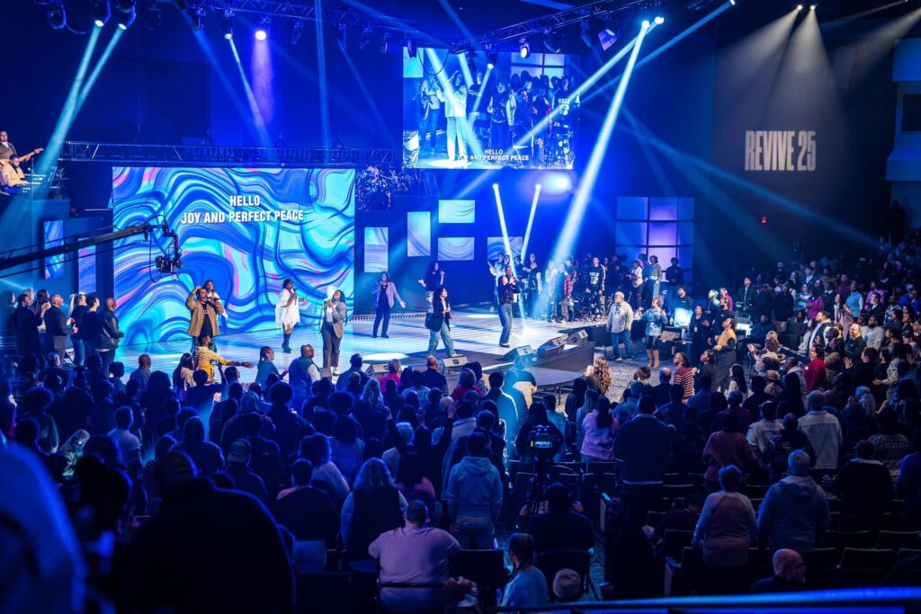 A vibrant worship service at Citygate Church with a large congregation gathered in a modern auditorium. The stage is illuminated with dynamic blue and purple lighting, featuring a live band and vocalists leading worship. A large LED screen displays the text "Hello, Joy and Perfect Peace," while additional lighting effects and a suspended camera crane enhance the immersive atmosphere. The crowd is engaged, standing and participating in the service, creating an energetic and communal environment.